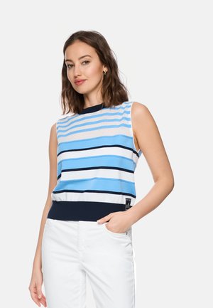 Frau trägt ärmelloses blau-weiß-schwarz gestreiftes Oberteil und weiße Hose, steht mit einer Hand in der Tasche vor einfachem Hintergrund.