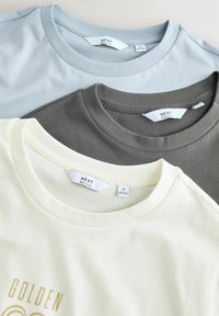 Tre crew-neck t-shirts: lyseblå, mørkegrå og creme. Hver har et mærke med teksten "NEXT" og en blød bomuldsstruktur.