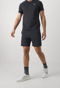 Černé atletické tričko a šortky, přiléhavý design, lehký materiál, s reflexním logem. Bílá sportovní obuv a šedé ponožky dotvářej í outfit.