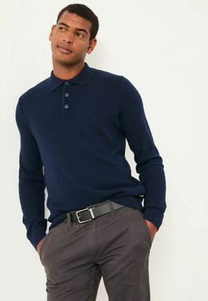 Marineblaues Longsleeve-Polo-Strickpullover mit Drei-Knopf-Blende, getragen über dunkelgrauen Hosen, ergänzt durch einen schwarzen Gürtel mit einer silbernen Schnalle.