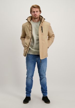 Beige jack met een capuchon en ritszakken, gedragen over een lichtgroene trui, gecombineerd met blauwe jeans en zwarte schoenen.