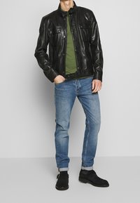 Man i svart läderjacka, olivgrön skjorta, blå jeans med uppvikta fållar och svarta spänneskor framför en enkel grå bakgrund.