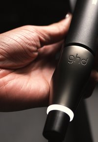 Czarny przyrząd do stylizacji włosów trzymany w ręce, z gładkim matowym wykończeniem, okrągłym białym paskiem świetlnym LED oraz wygrawerowanym logo „ghd”.