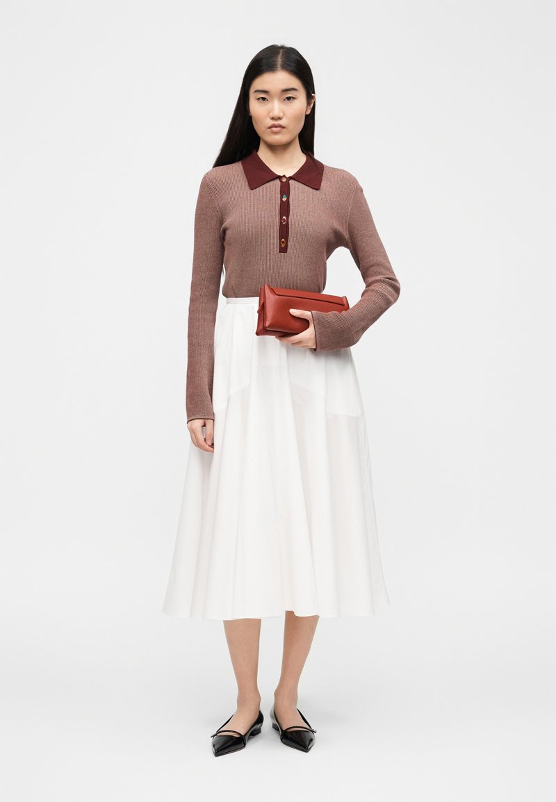 Donna in piedi che tiene una pochette color ruggine, indossando una maglietta marrone a maniche lunghe con colletto, una gonna midi bianca e ballerine nere con punta.