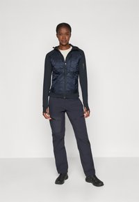 Veste matelassée marine avec fermeture éclair, devant texturé et manches longues, associée à un pantalon sombre avec poches zippées et coupe ajustée.