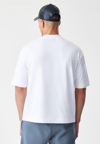 Bijela pamučna majica s kratkim rukavima, opuštenog kroja i okruglim izrezom. Glatka tekstura, minimalistički dizajn, bez vidljivih logotipa ili akcenta.