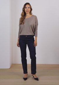 Beige strikket sweater med trekvartærmer, parret med mørkeblå jeans. Modellen står i sorte spidse hæle foran en neutral baggrund.