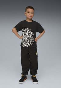 Junger Junge steht mit den Händen in den Hüften, trägt ein schwarzes Grafik-T-Shirt, passende schwarze Cargo-Hosen und schwarze Sneaker mit orangefarbigen Akzenten.