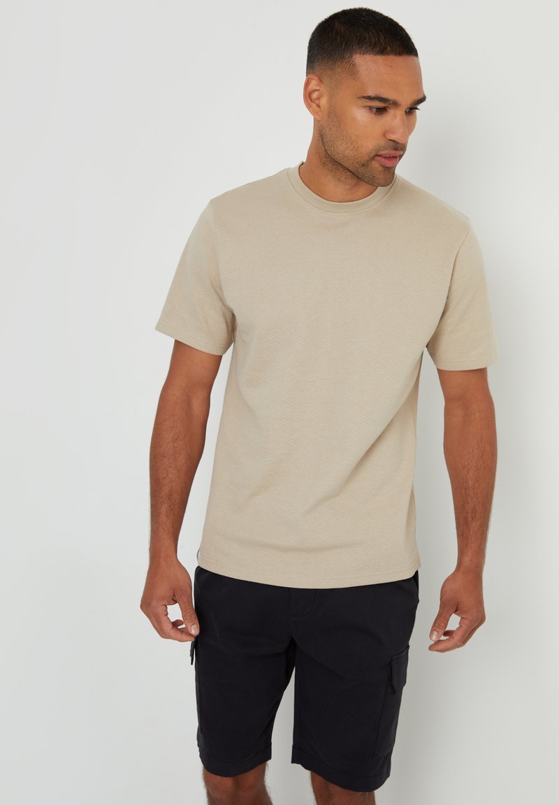 Beige texturované tričko s kulatým výstřihem a krátkými rukávy, zkombinované s tmavými cargo šortkami. Tričko má volný střih a jednoduché designové prvky.