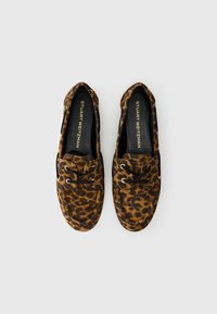 Un par de mocasines de ante con estampado de leopardo, con cordones negros y ojales dorados, etiquetados como Stuart Weitzman, mostrados desde arriba sobre un fondo blanco.