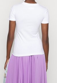 Biała bawełniana koszulka z krótkim rękawem, dopasowany krój, okrągły dekolt, noszona z plisowaną lawendową spódnicą, gładka tekstura.