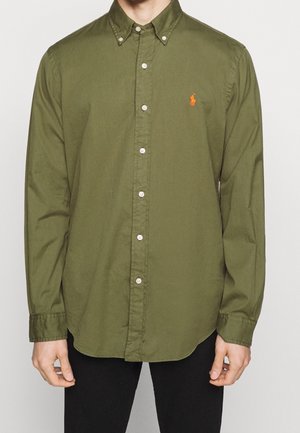 Mężczyzna ubrany w oliwkowo-zieloną koszulę z długim rękawem i guzikami, z haftowanym pomarańczowym logo na piersi, stojący na jednolitym tle.