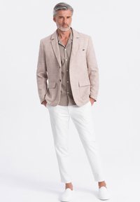 Jasno beżowa lniana marynarka z klapami, zestawiona z taupe'ową koszulą na guziki oraz białymi, slim-fit spodniami i butami typu slip-on.