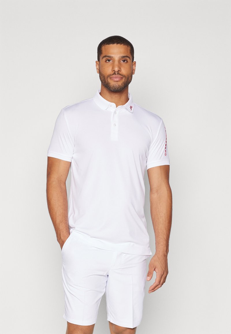 Vit polo skjorta med krage, korta ärmar och en treknappslåst placket. Har en röd logotyp på vänster axel. Bärs med vita shorts.