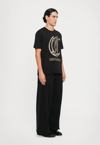 Μαύρο βαμβακερό t-shirt με χρυσό γραφικό σχέδιο και κείμενο, σε συνδυασμό με φαρδιά μαύρα παντελόνια και μαύρα παπούτσια, μοντέλο σε πλάγια στάση.