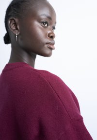 Jeune femme noire avec des cheveux tressés portant un pull rouge foncé, tournée vers la droite sur un fond blanc uni.