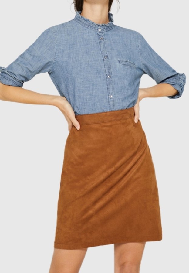 Camicia in denim con colletto arricciato abbinata a una mini gonna in suede marrone con design drappeggiato. Entrambi i pezzi sono aderenti.