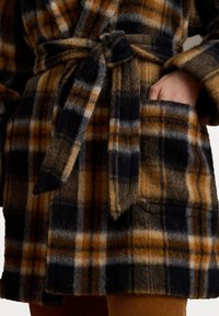 Accappatoio in lana a quadri nei toni del blu scuro, arancione e crema; presenta una cintura da annodare, tasche laterali e un tessuto morbido e texturizzato.