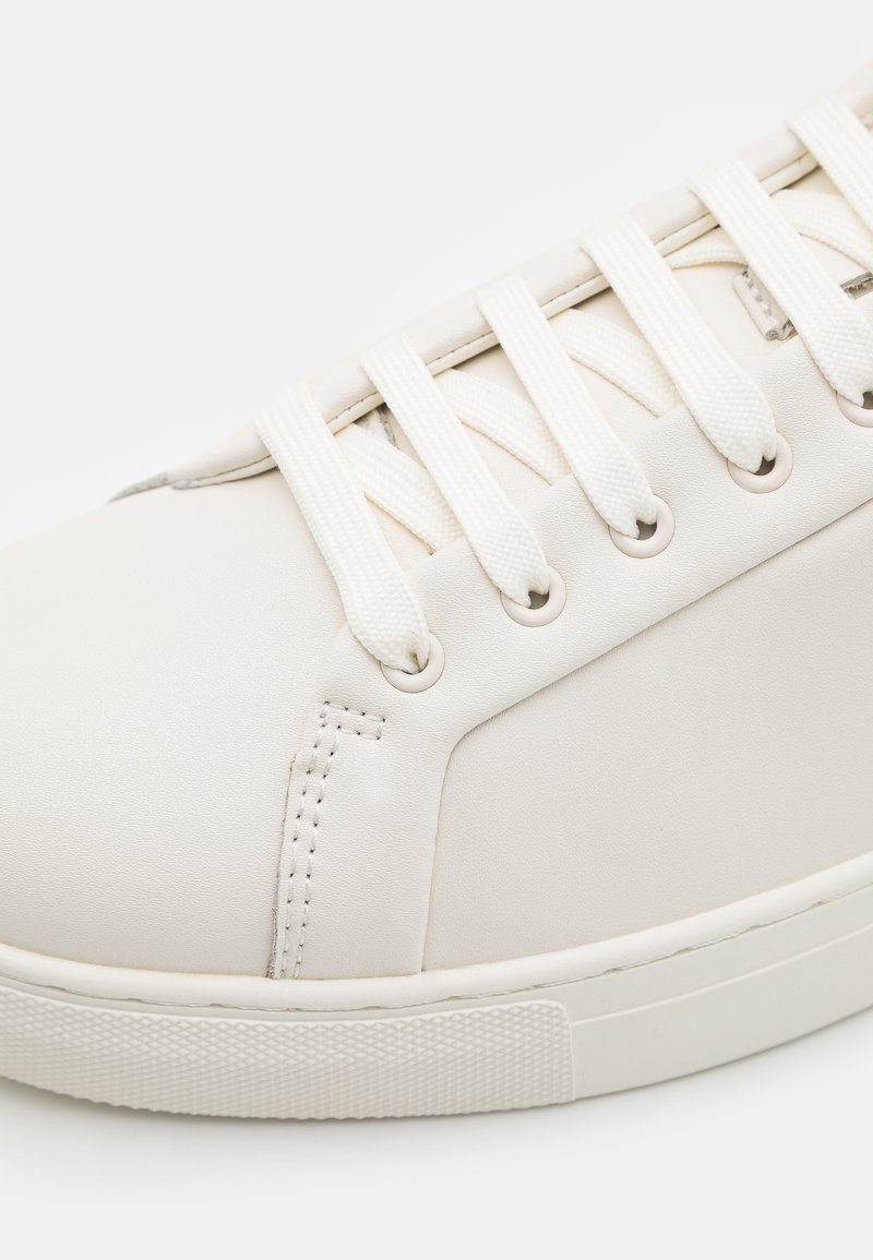 Sneaker en cuir blanc avec une texture lisse, featuring des lacets plats blancs, des œillets, et une semelle en caoutchouc avec un motif de grip texturé.