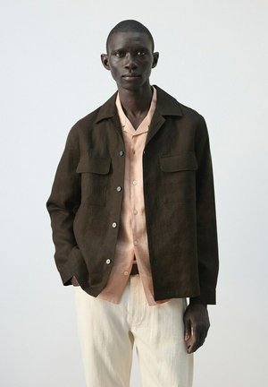 Hombre con chaqueta marrón oscuro abotonada sobre una camisa color durazno claro y pantalones crema, de pie frente a un fondo liso y claro.