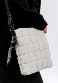 Borsa grigia in ecopelle trapuntata con forma rettangolare, dotata di una tracolla intrecciata e hardware nero. Appoggiata su un outfit nero.