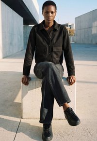 Ung person med kort hår iført sort jakke, mørke jeans og sorte sneakers sidder med korslagte ben på en betonblok udendørs.