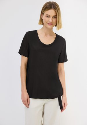 Svart kortärmad t-shirt med rundad halsringning och sidoknytning. Slätt tyg med en avslappnad passform. Kombinerad med ljusa vida byxor.