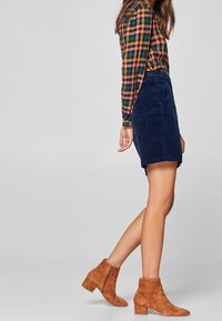 Jupe en velours côtelé marine associée à une chemise à carreaux multicolore et des bottines en suède marron avec un talon épais. Comprend des manches longues et une fermeture éclair.