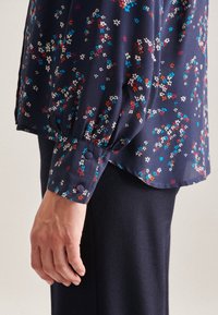 Blusa floreale blu navy con maniche a sbuffo, caratterizzata da piccoli fiori multicolori e polsini con bottoni. Tessuto morbido e leggero con una consistenza liscia.