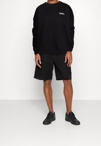 Hombre vistiendo un sudadera negra de manga larga con el texto "Offline", pantalones cortos negros hasta la rodilla y zapatillas negras, de pie sobre un suelo blanco.