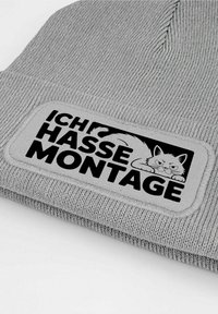 Gray knit beanie featuring a rectangular patch with a black cat illustration and the text "ICH HASSE MONTAGE" in bold black letters.