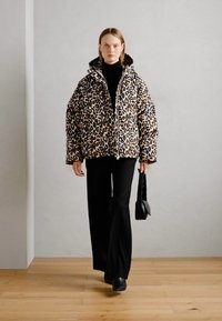 Chaqueta inflable con estampado de leopardo, cuello alto, cierre frontal y ajuste oversize, combinada con pantalones negros y un bolso pequeño. Fondo de suelo de madera.