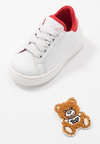 Baskets blanches avec doublure intérieure rouge et lacets blancs à côté d'un petit patch ours en peluche marron avec "TOY" écrit sur son t-shirt.