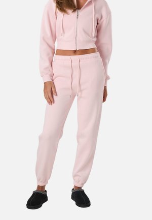 Kobieta ubrana w jasnoróżową krótką bluzę na zamek i pasujące spodnie dresowe, stojąca na białym tle, w czarnych butach na ślizg.