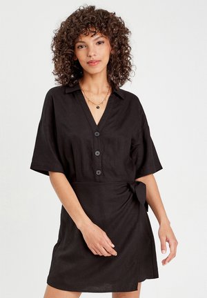 Robe noire en lin à manches courtes, décolleté en V, devant à boutons, avec un détail de lien sur le côté à la taille. Texture douce et coupe décontractée.