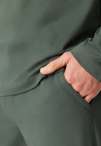 Top de tela verde con textura que cuenta con un bolsillo lateral. Vista en primer plano de una mano descansando en el bolsillo. Detalles de costura sutiles visibles.