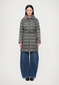 Hall hall talvejope karusnahast krae, vöötatud vöökoht, pikad varrukad ja esikinnis tõmblukuga; kantud laiade säärega teksapükste ja mustade kingadega.