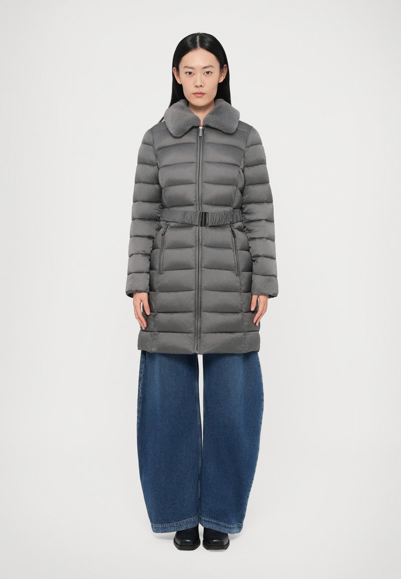 Hall hall talvejope karusnahast krae, vöötatud vöökoht, pikad varrukad ja esikinnis tõmblukuga; kantud laiade säärega teksapükste ja mustade kingadega.