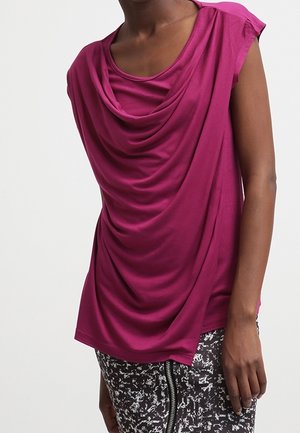 Nainen, joka pukeutuu hihattomaan magentanväriseen liehuvaan yläosaan ja mustavalkoiseen kuvioituun hameeseen, jossa on etuvetoketju, seisoo yksiväristä taustaa vasten.