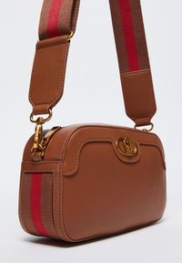 Sac à bandoulière en cuir marron avec un fermoir en or, présentant une surface texturée et une bandoulière en tissu rayé dans des tons marron foncé et rouge.