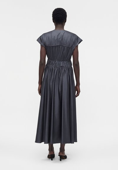 Μαύρο μακρύ φόρεμα χωρίς μανίκια με σούρες στη μέση και στους ώμους, φορεμένο με μαύρα σανδάλια με τακούνι, μοντέλο με πλάτη σε λευκό φόντο.