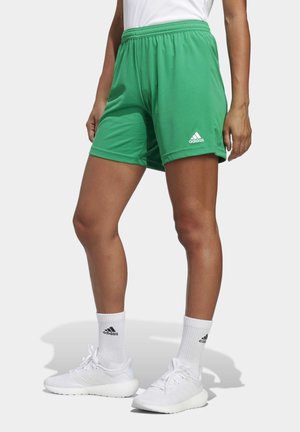 Osoba nosząca zielone szorty Adidas, białe skarpetki Adidas i białe buty sportowe, stojąca na jednolitym tle.