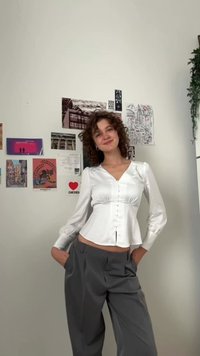Bijela satenska majica s V-izrezom, dugim napuhanim rukavima i gumbima sprijeda, u kombinaciji s hlačama visokog struka u sivoj boji koje imaju pliće i bočne džepove.