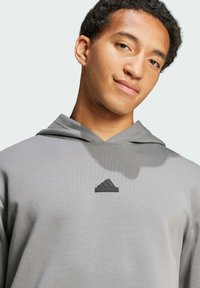 Světle šedá mikina vyrobená z měkké látky, s klokaní kapsou a černým logem Adidas na prsou. Má kapuci se stahovací šňůrkou.