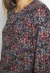 Blommig blus med mörk bakgrund och röda, blå och krämfärgade blommor. Inkluderar plisserad detalj vid halsringningen. Mjuk och lätt vikt.
