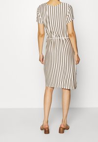 Robe beige à manches courtes, longueur genoux, avec des rayures verticales brunes. Présente une taille cintrée et une texture subtile. Une paire de sandales à talons brunes.