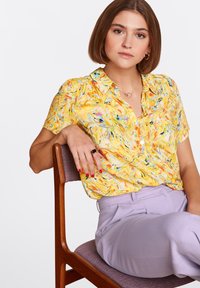 Żółta bluzka z krótkim rękawem w wirującym wzorze; zestawiona z jasnofioletowymi spodniami. Modelka siedzi na drewnianym krześle.