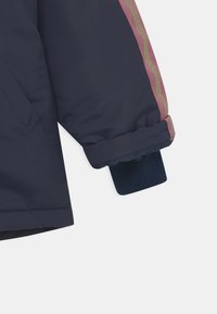 Manche de veste bleu marine avec un revers plié, présentant une bande ondulée réfléchissante rose et dorée le long du bras externe.