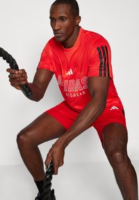 Punainen Adidas-urheiluasu sisältää teksturoitua jerseykangasta ja shortseja, joissa on mustat kolme raita -korostukset. Malli pitää mustasta treenirihmasta kiinni.