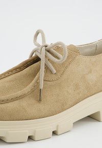 Gros plan sur une chaussure en daim beige avec une épaisse semelle crême et des lacets ronds beiges noués, mettant en valeur les détails cousus sur la tige.